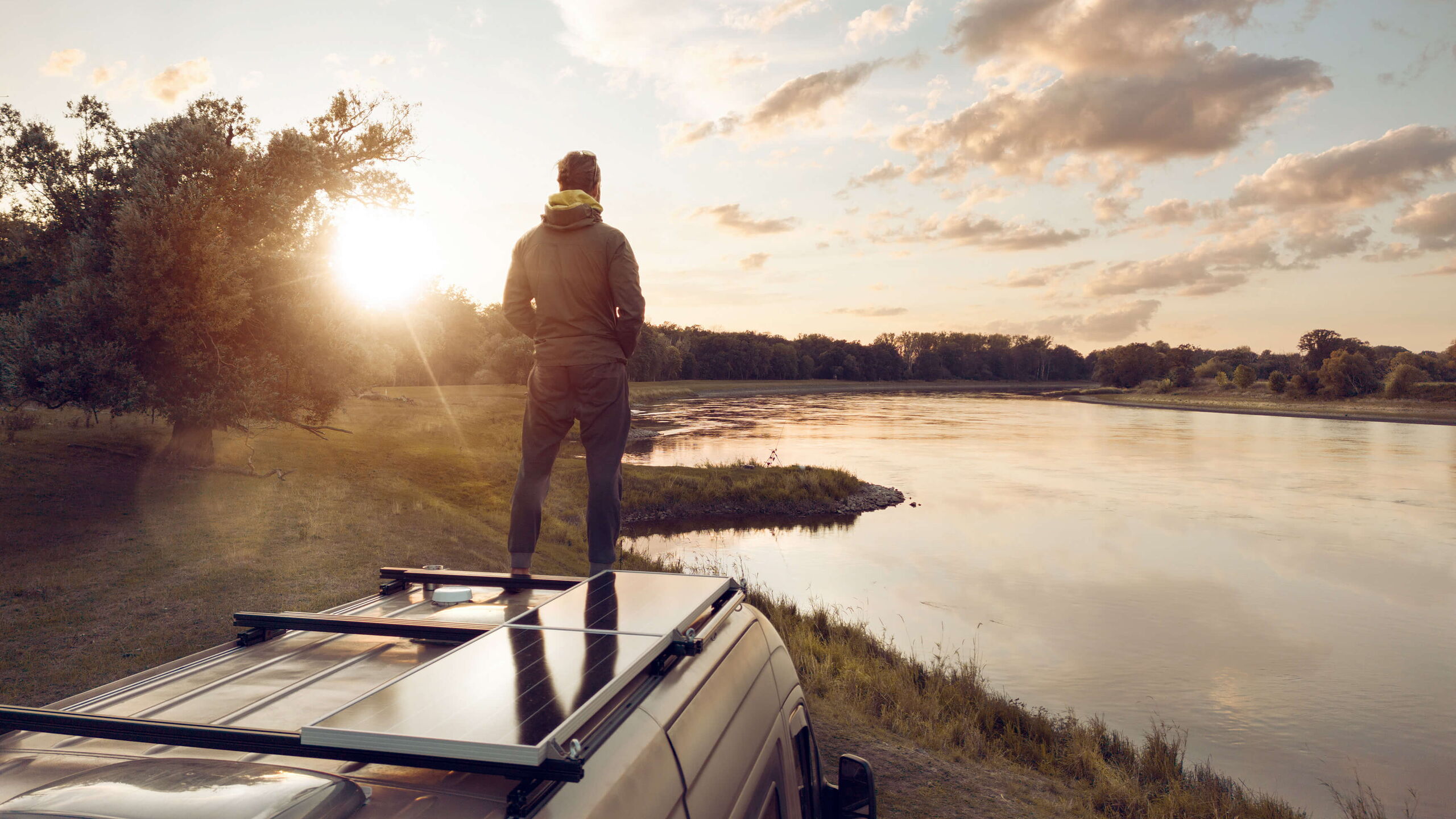 Unabhängig ist klug. Ein Mann steht auf dem Dach seines Campers und schaut einen Fluss entlang in den Sonnenuntergang.